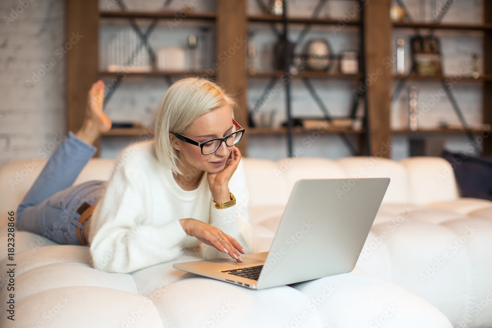 Fototapeta premium Pretty young blonde girl working on a laptop sitting on the bed. Beautiful young woman in a fluffy white sweater and glasses working from home on a laptop lying on the bed in the room with white walls