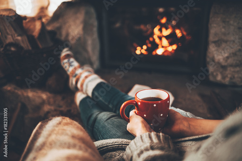 Woman in woollen socks by the fireplace. Unrecognisable relaxes by warm fire with a cup of hot drink and interesting book, warming up her feet. Cozy atmosphere. Winter and Christmas holidays concept. photo