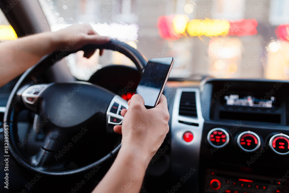 Driver with a smartphone in hand in the driver's seat. The concept of inattention at the wheel. Driving with one hand, distracted by SMS or call by mobile. Cropped hand of businessman using phone