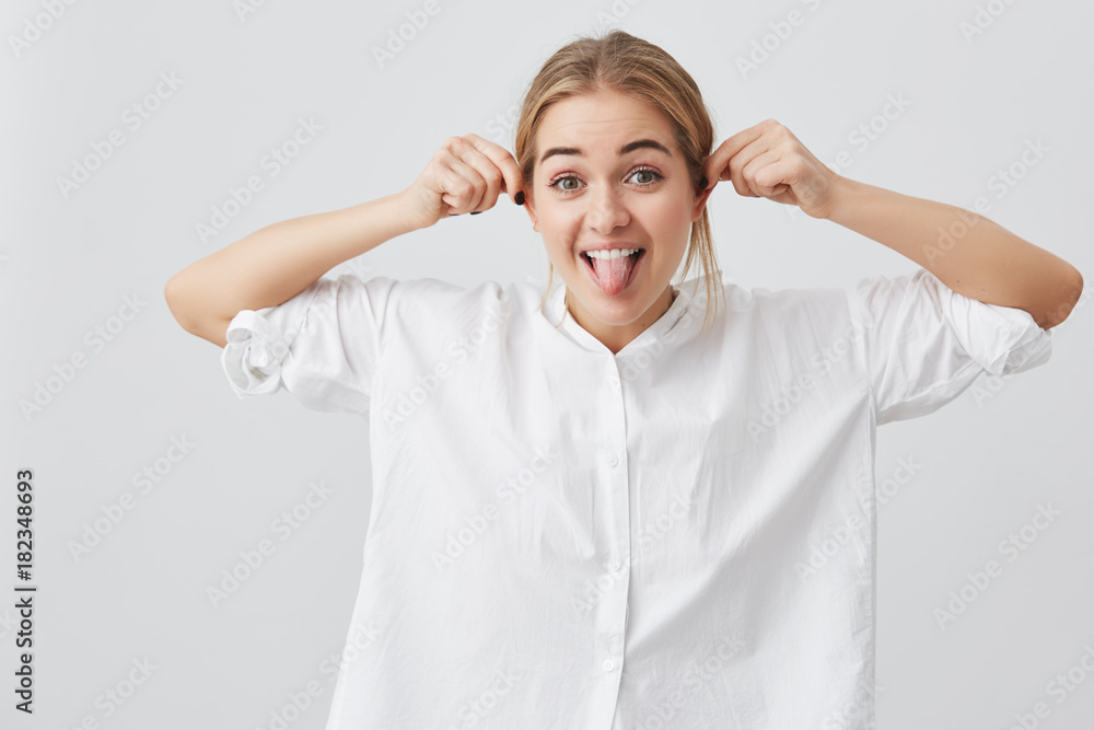 Playful funny pretty girl with gloomy expression demonstaring tongue, pulling herself at ears, mocking at camera. Playful beautiful european female with fair hair posing against gray background. Playful funny pretty girl with gloomy expression demonstaring tongue, pulling herself at ears, mocking at camera. Playful beautiful european female with fair hair posing against gray background.