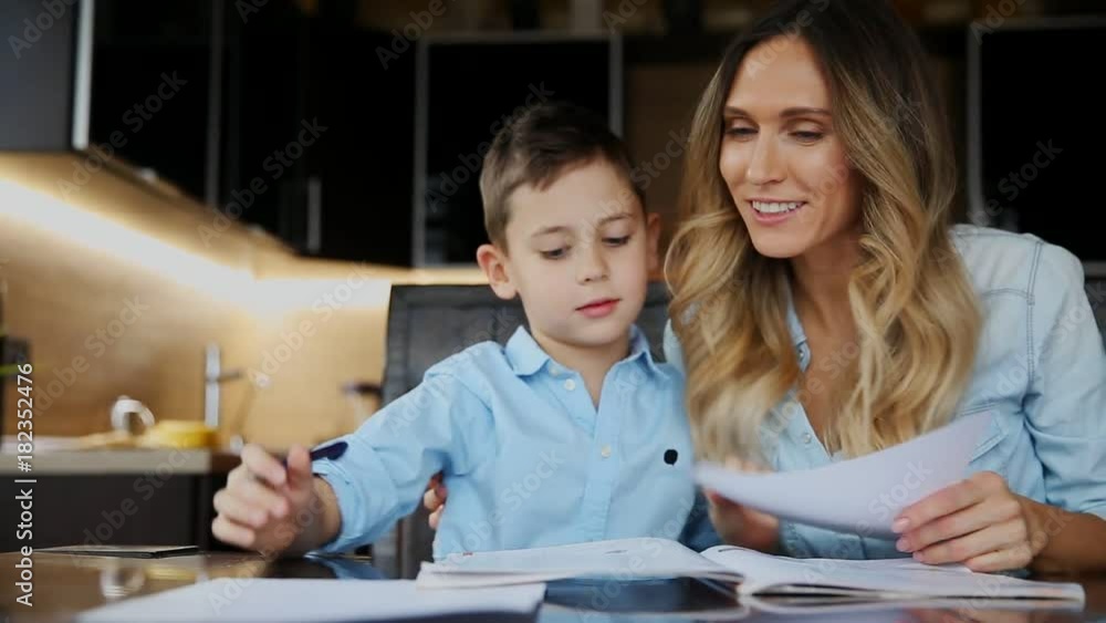 Smiling mother helping his son doing homework, dictates the text to help you write sitting at the table in the kitchen. Smiling mother helping his son doing homework, dictates the text to help you write sitting at the table in the kitchen.
