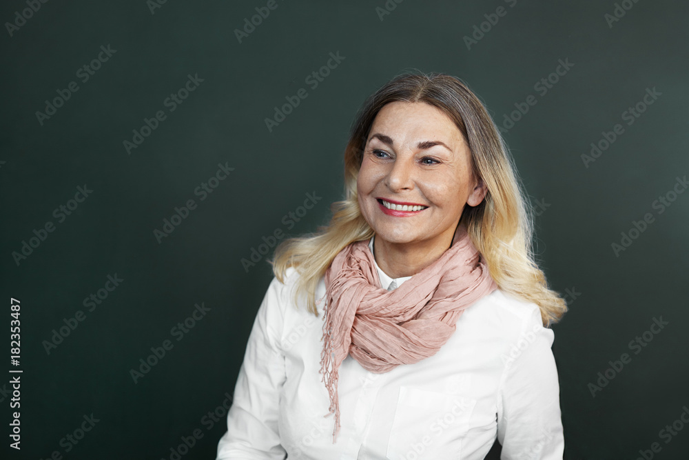 Obraz premium Picture of elegant beautiful elderly 60 year old woman with blonde loose hair and charming smile posing in studio, looking sideways with interest, smiling, showing her perfect white healthy teeth