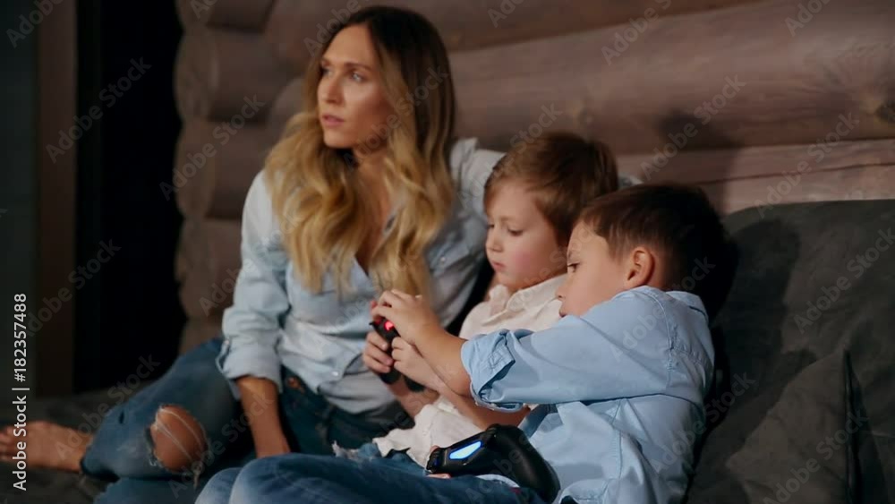 Mother and two sons sitting on the sofa in his house playing video games with wireless joystick. Happy people in the house. Mother and two sons sitting on the sofa in his house playing video games with wireless joystick. Happy people in the house.