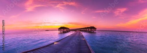 Sunset on Maldives island, water villas resort. Beautiful sky and clouds. Beautiful beach background for summer travel with sun, beach wooden jetty. Summer mood sun beach background concept.