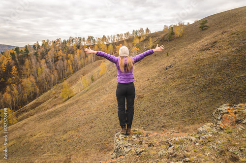 A girl in a lilac jacket stretching her arms on a mountain, a view of the mountains and an autumn forest by a cloudy day, free space for text
