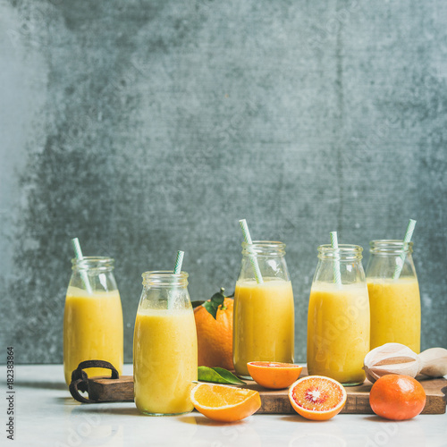 Healthy yellow smoothie with citrus fruit and ginger in bottles on wooden board over light marble table, selective focus, copy space, square crop. Clean eating, vegan, dieting food concept