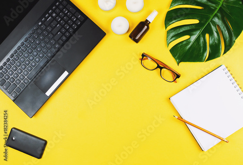 Business flatlay with laptop, mobile phone, glasses, philodendron leaf, candles, cream, pen and notebook. Concept of a woman's work place. Flat lay. Yellow background.