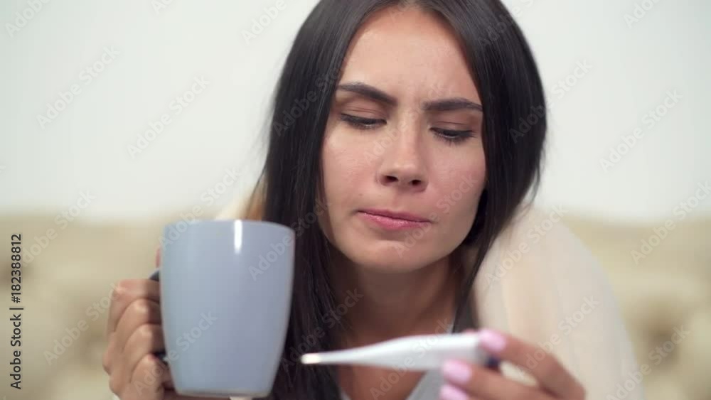 So high. Sick young woman drinking tea and holding a thermometer while sitting on the sofa So high. Sick young woman drinking tea and holding a thermometer while sitting on the sofa