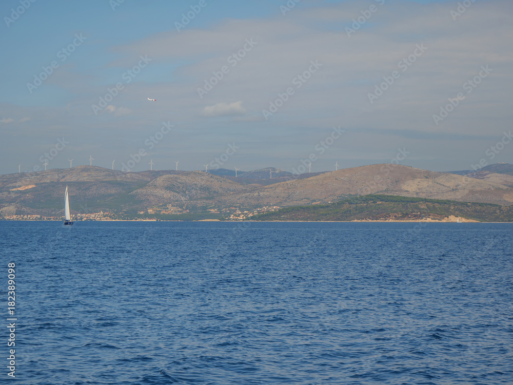 Croatian coastline with sailing boat and wind power generators on hills.