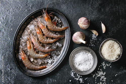Raw whole fresh uncooked prawns shrimps on ice on vintage metal tray with cooking ingredients above. Sea salt, rice, garlic, over black texture background. Top view with space