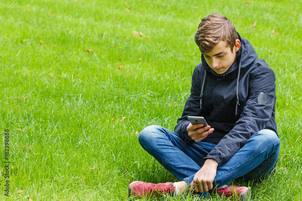 Male student talking on a phone seated on a grass in the city park