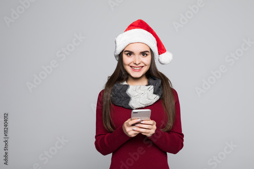 Portrait of a brunette young woman in red santa claus using mobile phone isolated on the gray background