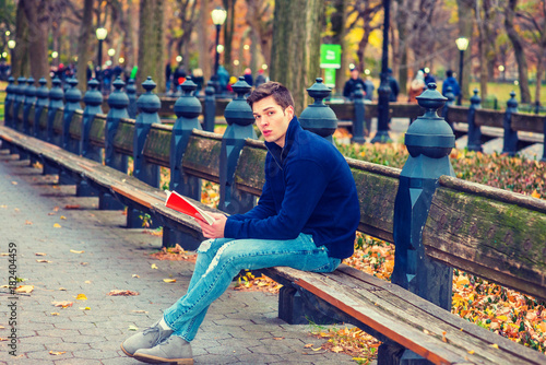 Young American man wearing blue sweatshirt, fashionable destroyed jeans, gray casual shoes, sits on long bench at Central Park, New York in autumn day, looks down, reads red book. Filtered effect..