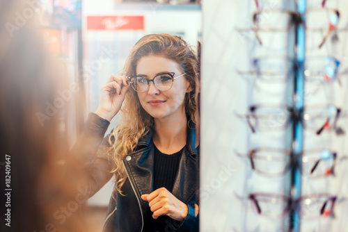 Smiling young woman trying on glasses on mirror in optician.