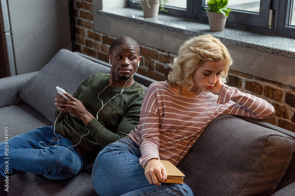 Obraz premium african american man in earphones looking at sad girlfriend with book on sofa at home