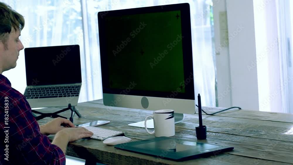 A caucasian male freelance designer, programer working at Home with all in one unit computer with green screen, laptop, a pen tablet and other electronics peripheral on the wooden table on a bright mo A caucasian male freelance designer, programer working at Home with all in one unit computer with green screen, laptop, a pen tablet and other electronics peripheral on the wooden table on a bright mo