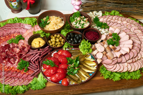 Top view shot of a huge wooden plate full of sliced meat appetizers salami ham different kinds of sausages served with mustard olives chopped mushrooms tomatoes and sliced cucumbers.