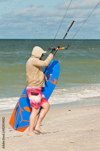 Young male walking, tropical, sandy beach carrying a kiteboard, holding the handlebar of a cabled canopy on a windy day, shoreline on the Gulf of Mexico