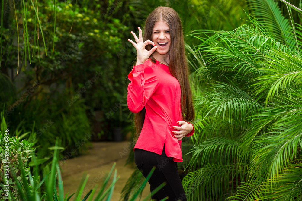 Young happy women shows the OK symbol by hand in a red blouse standing in the palm garden Young happy women shows the OK symbol by hand in a red blouse standing in the palm garden