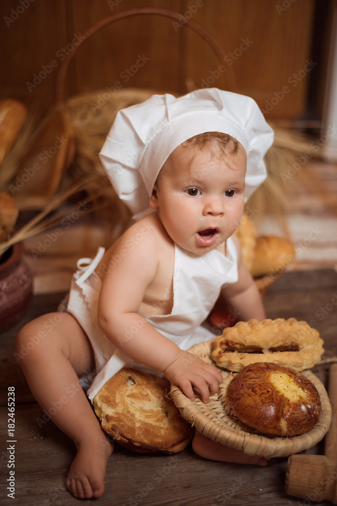 little boy in a cook costume among a baking bread rolls of flour confectionery on a brown wooden floor sits on a wooden background little boy in a cook costume among a baking bread rolls of flour confectionery on a brown wooden floor sits on a wooden background