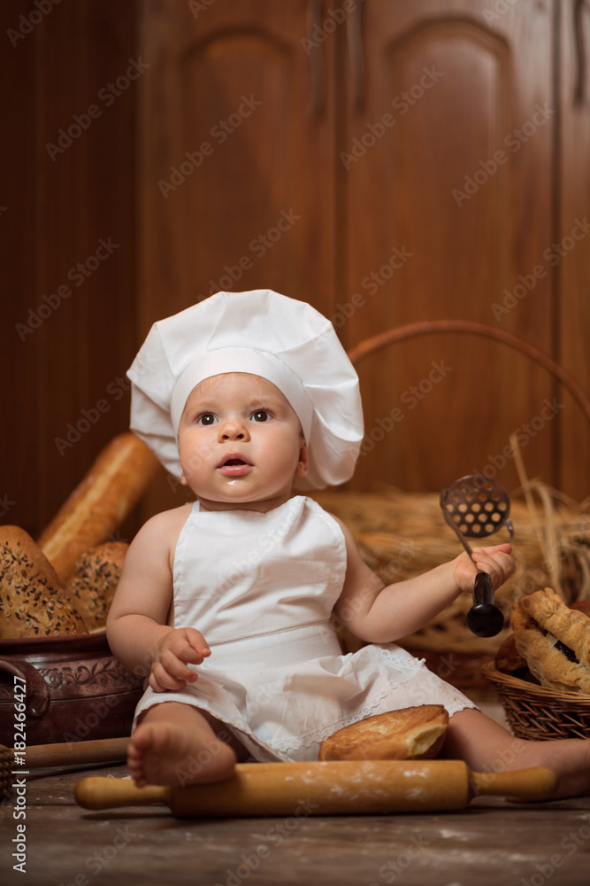 Happy little child in the cook costume at the kitchen sitting on the table among baskets with tasty rolls, buns and bread. Copy space Happy little child in the cook costume at the kitchen sitting on the table among baskets with tasty rolls, buns and bread. Copy space