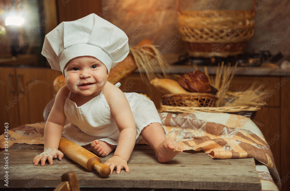 Happy little child in the cook costume at the kitchen sitting on the table among baskets with tasty rolls, buns and bread. Copy space Happy little child in the cook costume at the kitchen sitting on the table among baskets with tasty rolls, buns and bread. Copy space