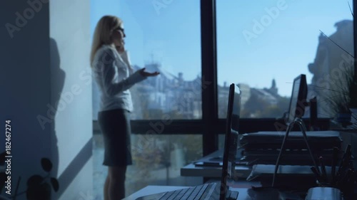 Slow motion footage of beautiful blonde businesswoman talking on the mobile phone and looking over city view from office window / Businesswoman talking on the mobile phone