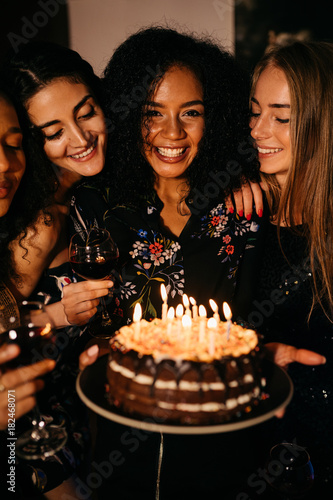 Canvas Print Young happy woman holding a birthday cake, standing indoors surrounded by girlfr