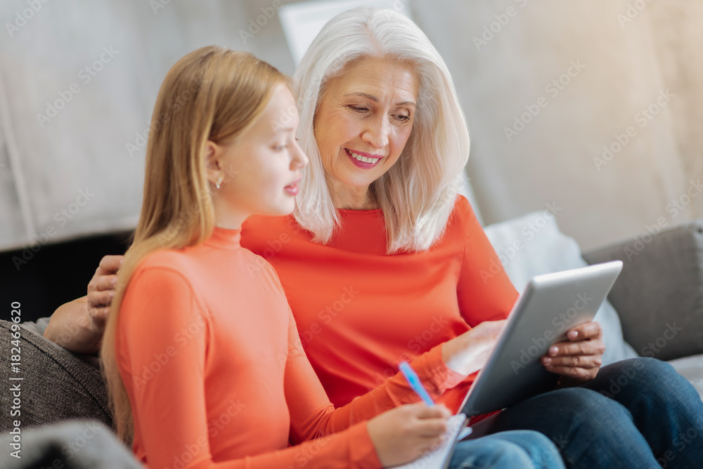 What is this. Smart nice curious girl pointing at the tablet screen and asking her grandmother a question while sitting with her on the sofa What is this. Smart nice curious girl pointing at the tablet screen and asking her grandmother a question while sitting with her on the sofa