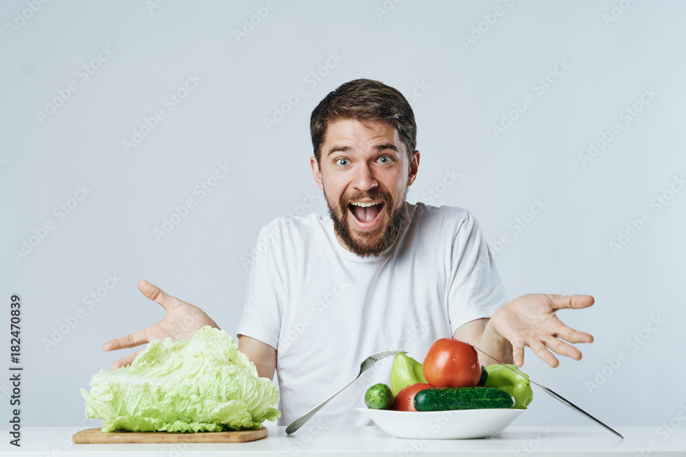 Man with a beard on a light background sits at a table in front of a plate full of vegetables, diet, vegetarian Man with a beard on a light background sits at a table in front of a plate full of vegetables, diet, vegetarian