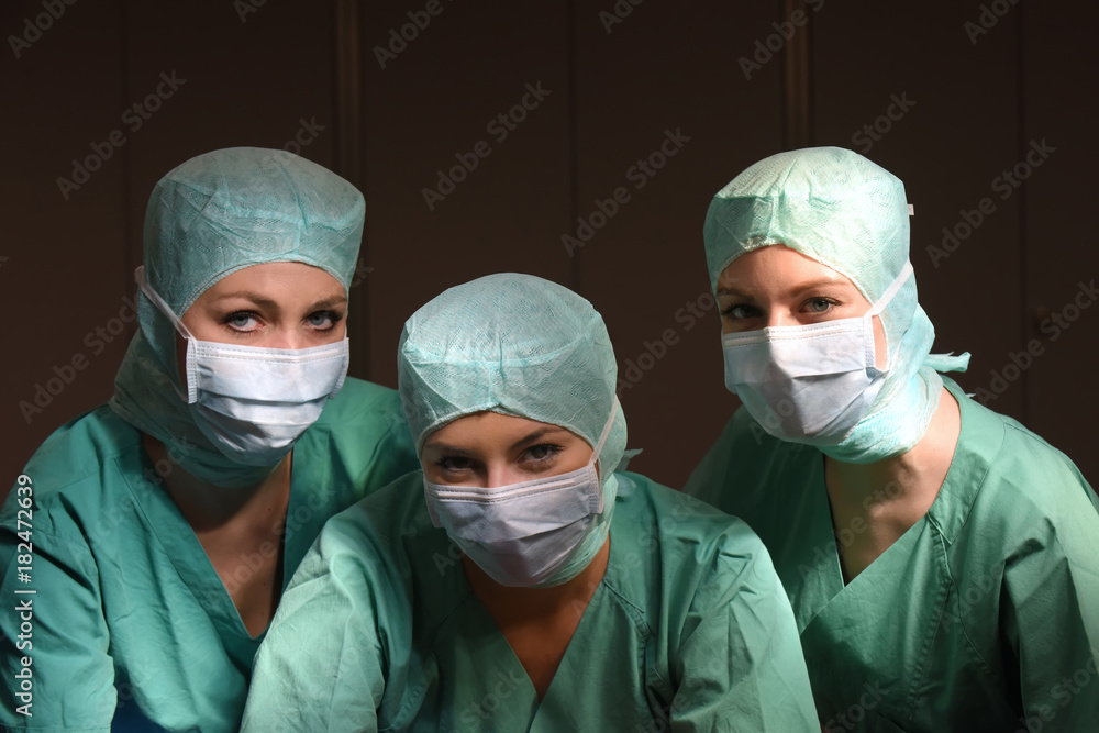 A group of three young women pose in a medical operation theater. Fully dressed as theater nurses with face masks and green sterile medical work clothing. A group of three young women pose in a medical operation theater. Fully dressed as theater nurses with face masks and green sterile medical work clothing.