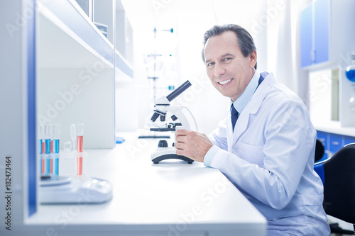 In the lab. Happy positive professional scientist sitting at the table and using a microscope while working in the lab