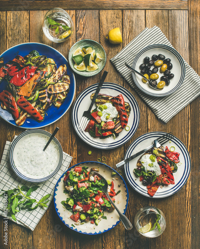 Flat-lay of healthy dinner table setting. Fresh salad, grilled vegetables with yogurt sauce, pickled olives, lemon water over wooden background, top view, square crop. Clean eating, vegetarian food