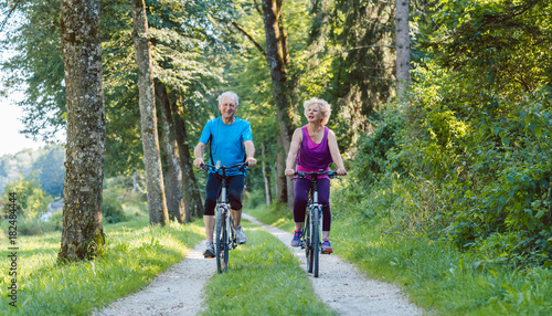 Full length of a happy and active senior couple wearing cool fitness outfits while riding bicycles outdoors in the park