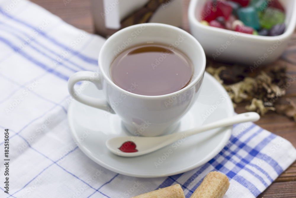 Still life with a white porcelian tea cup,saucer and spoon on a white blue plaid napkin and a bowl of colored candies on a wooden table Still life with a white porcelian tea cup,saucer and spoon on a white blue plaid napkin and a bowl of colored candies on a wooden table