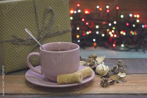 Festive still life with a pink porcelian cup of tea,a saucer, a teaspoon, cookies, a golden giftbox and fairy lights, colored wooden background photo