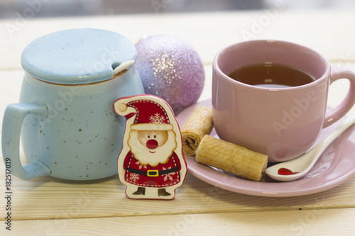 A festive still life with a pink porcelian cup of black tea, a saucer, a white spoon, a light blue closed sugar bowl and Cristmas-tree toys on a light wooden background photo