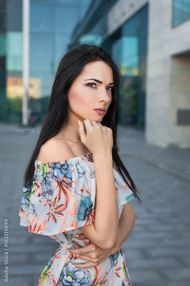 Young thoughtfull beautiful brunette woman with long hair in the street wearing white dress with flower pattern on the background of skyscraper