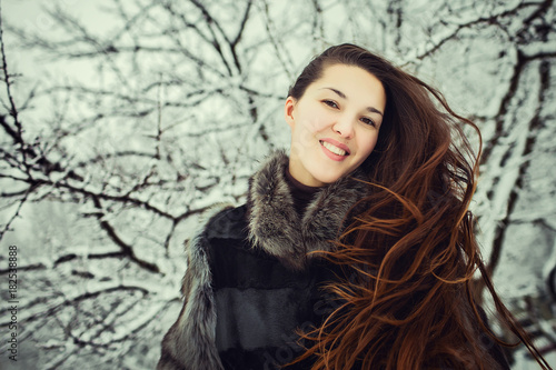portrait of beautiful young woman with long brunette hair in fur coat smiling and spending time outdoors in the winter forest.