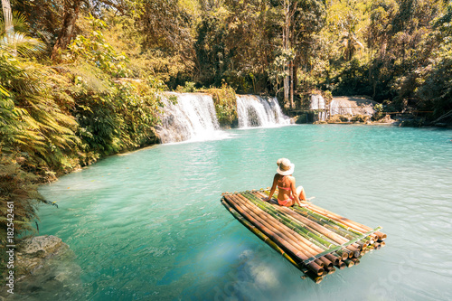 Woman in bikini and hat sitting on bamboo raft and enjoying view on waterfall. photo