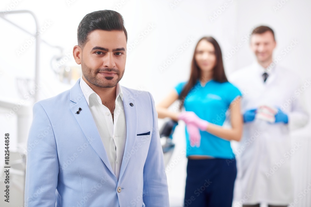 Handsome young man smiling to the camera posing at the dental clinic his dentist and nurse standing on the background copyspace professionalism trust happiness service dentistry oral smile. Handsome young man smiling to the camera posing at the dental clinic his dentist and nurse standing on the background copyspace professionalism trust happiness service dentistry oral smile.