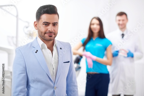 Handsome young man smiling to the camera posing at the dental clinic his dentist and nurse standing on the background copyspace professionalism trust happiness service dentistry oral smile.