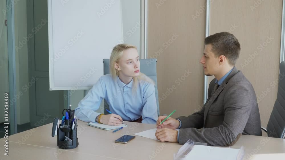 The young female student has a consultation with her male supervisor in his office. People sit at the big wooden table in the comfortable chairs and taking some notes with their pens. The professor in The young female student has a consultation with her male supervisor in his office. People sit at the big wooden table in the comfortable chairs and taking some notes with their pens. The professor in