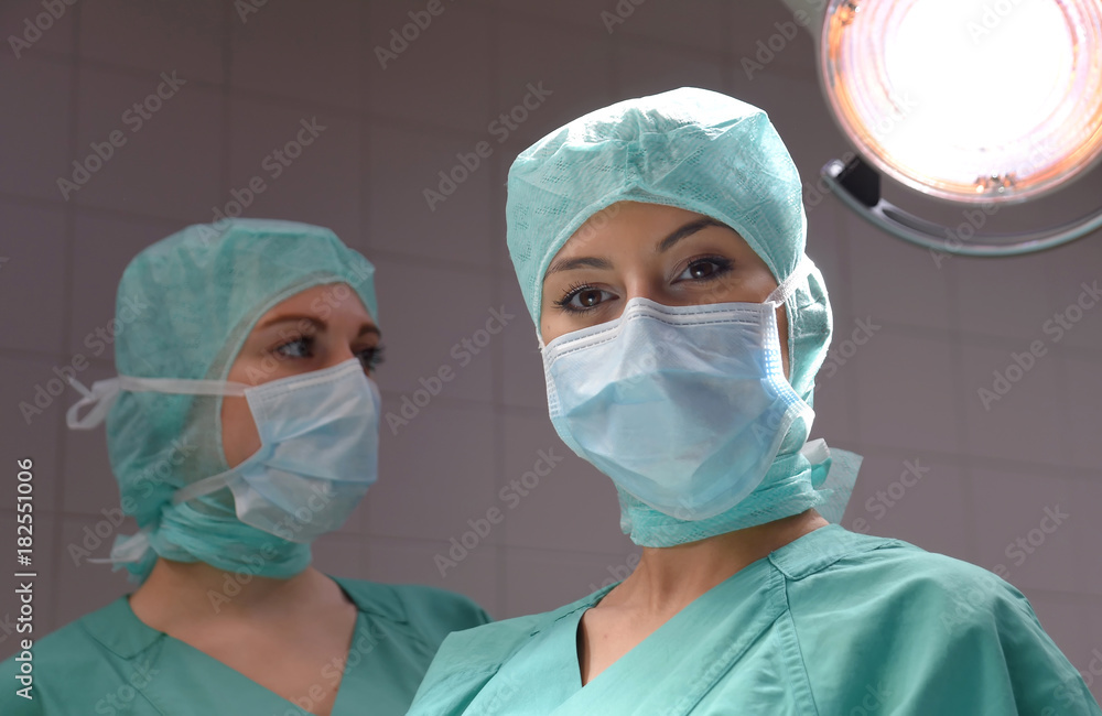Two young women pose in a low lit operation theater. Fully dressed as theater nurses with face masks and green sterile medical work clothing. Two young women pose in a low lit operation theater. Fully dressed as theater nurses with face masks and green sterile medical work clothing.