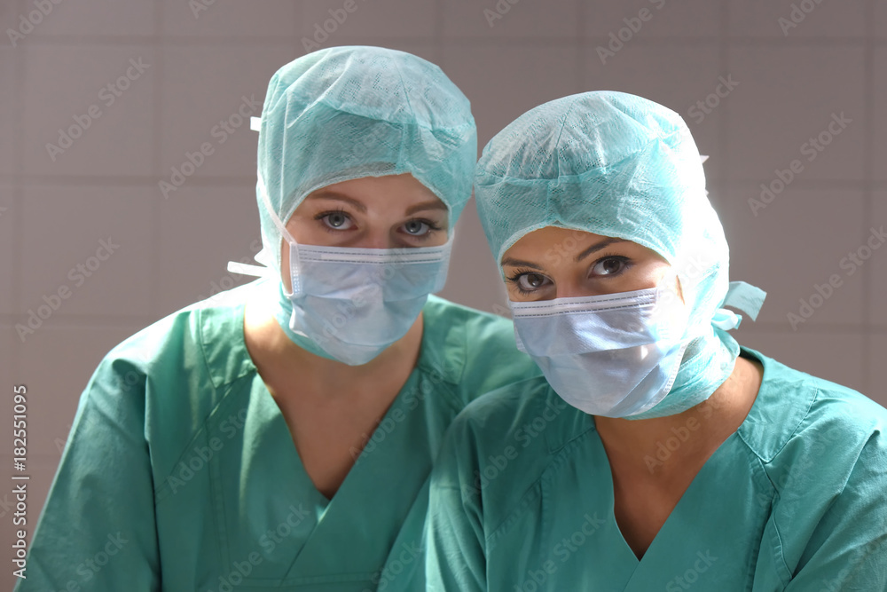 Two young women pose in a low lit operation theater. Fully dressed as theater nurses with face masks and green sterile medical work clothing. Two young women pose in a low lit operation theater. Fully dressed as theater nurses with face masks and green sterile medical work clothing.