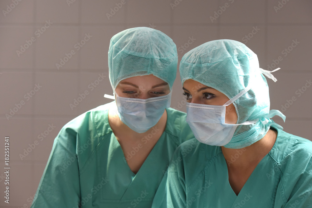 Two young women pose in a low lit operation theater. Fully dressed as theater nurses with face masks and green sterile medical work clothing. Two young women pose in a low lit operation theater. Fully dressed as theater nurses with face masks and green sterile medical work clothing.