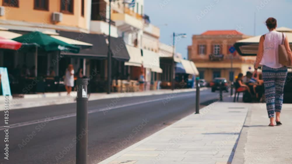 Young girl is walking along the sidewalk in a resort town in Europe on a bright sunny day. Cafes and restaurants in the background, cars and scooters pass by on the road. Ermoupolis city, Syros island Young girl is walking along the sidewalk in a resort town in Europe on a bright sunny day. Cafes and restaurants in the background, cars and scooters pass by on the road. Ermoupolis city, Syros island