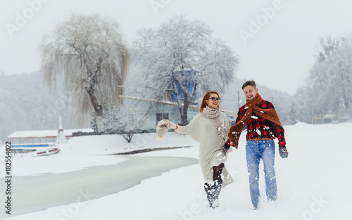 Full-length portrait of the beautiful couple holding hands while happily walking along the village covered with snow during the snowfall.