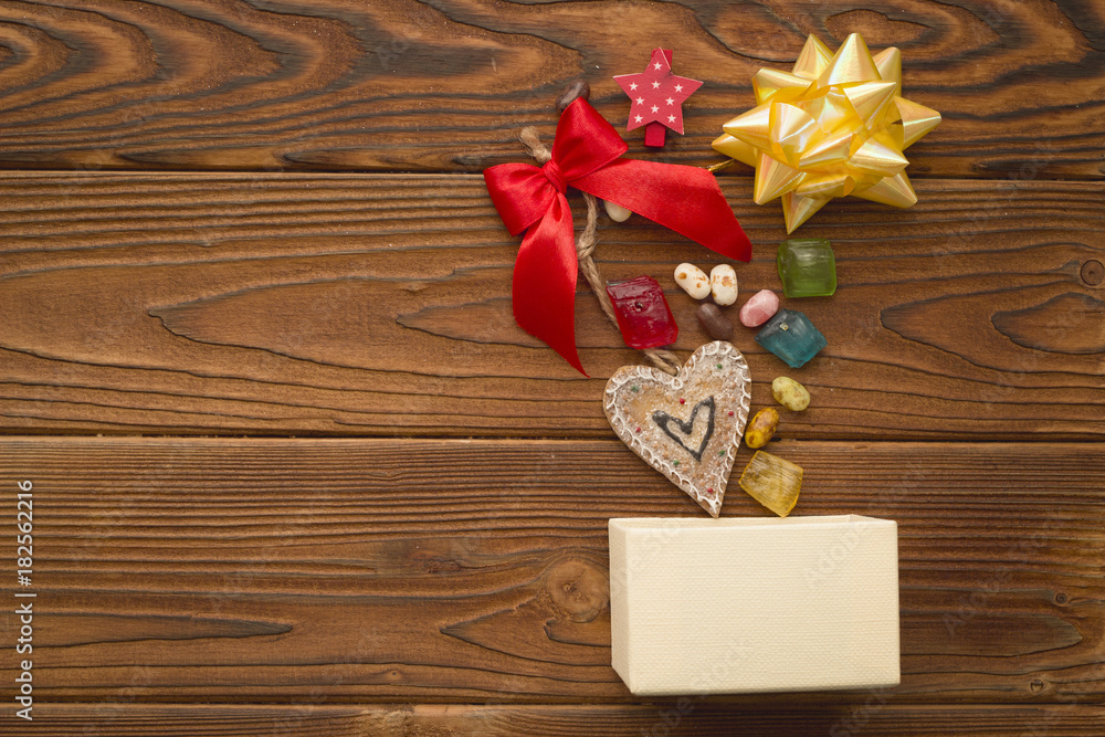 A beige box,a heart-shaped gingerbread, a red bow, a gold metallic bow and colored candies on a rustic wooden table. Christmas background. Copy space. Top view A beige box,a heart-shaped gingerbread, a red bow, a gold metallic bow and colored candies on a rustic wooden table. Christmas background. Copy space. Top view