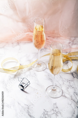 Two flute glasses with champagne on marble background. Pink transparent fabric. Soft light